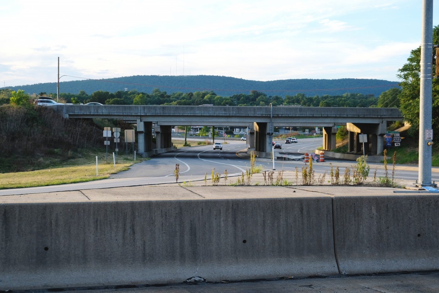 An intersection of a roadway with a concrete barrier in the foreground and a concrete bridge in the background. Trees and a low mountain range can be seen in the distance