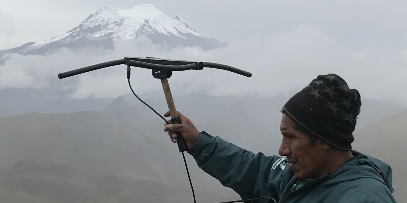 A researcher in Ecuador holds a transceiver up toward a tall, snowcapped mountain obscured by fog and clouds