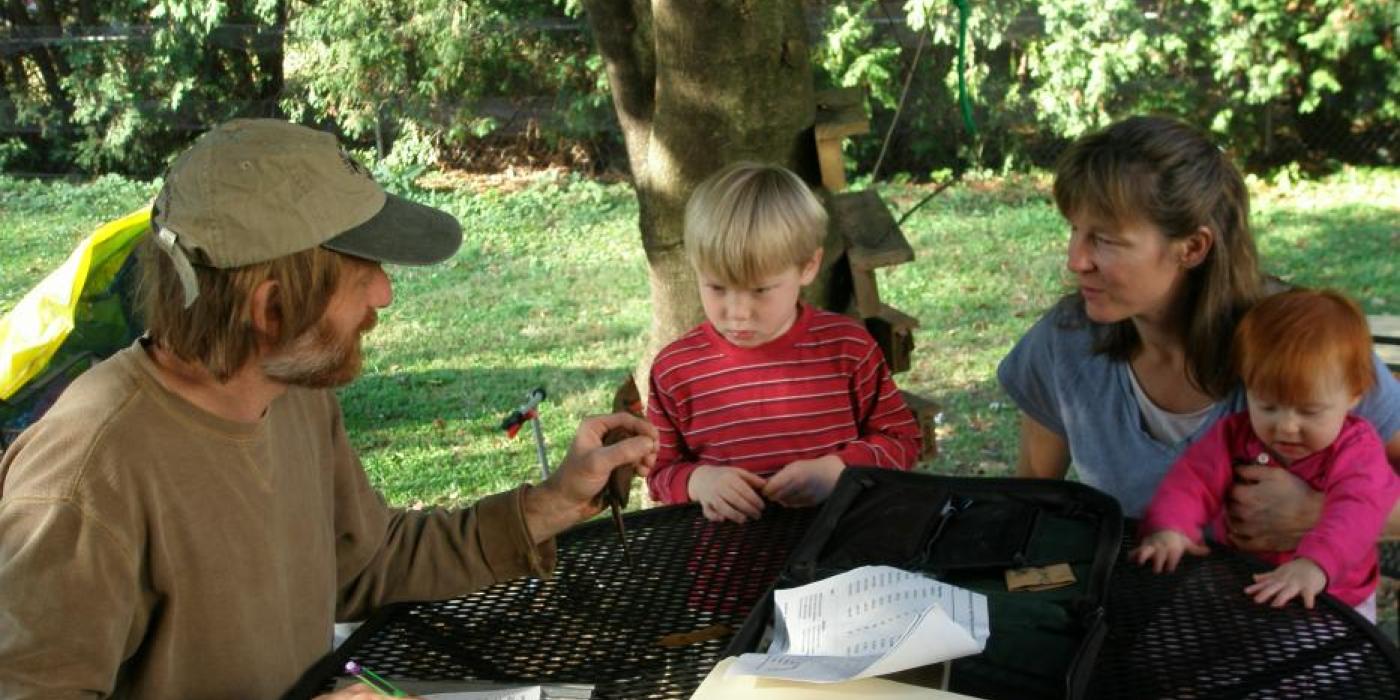 A researcher with the Smithsonian Migratory Bird Center talks to a mother and two young children about birds. They are seated around a table in their backyard with a tree and grass in the background.