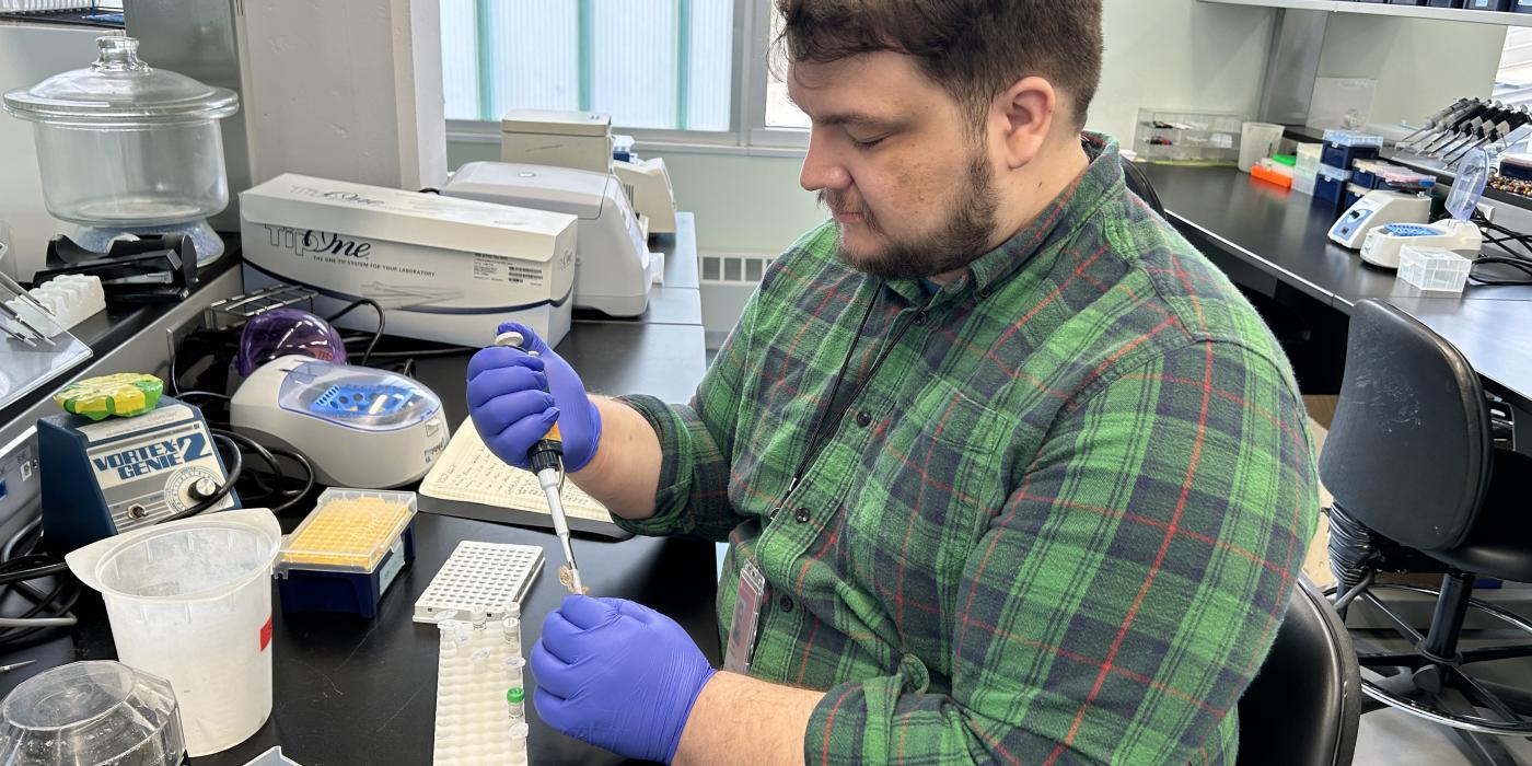 A photo of Sean Lyons in a lab setting. He is using a pipette to put fluid into a small vial. 