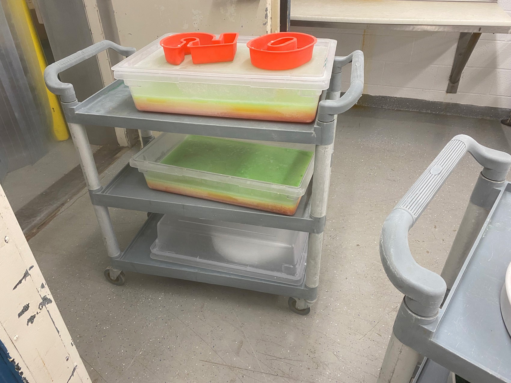 A three-tiered cart with rectangular plastic bins and a silicone number "50" mold filled with colorful frozen water and diluted fruit juice