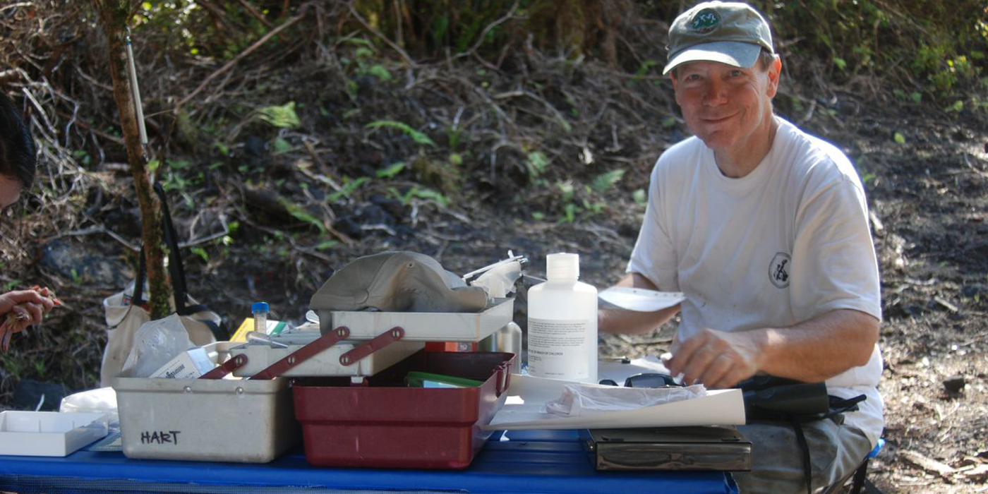 Portrait of a man wearing a white t-shirt and a blue baseball cap sits at a desk filled with scientific equipment in the Hawaiian jungle.