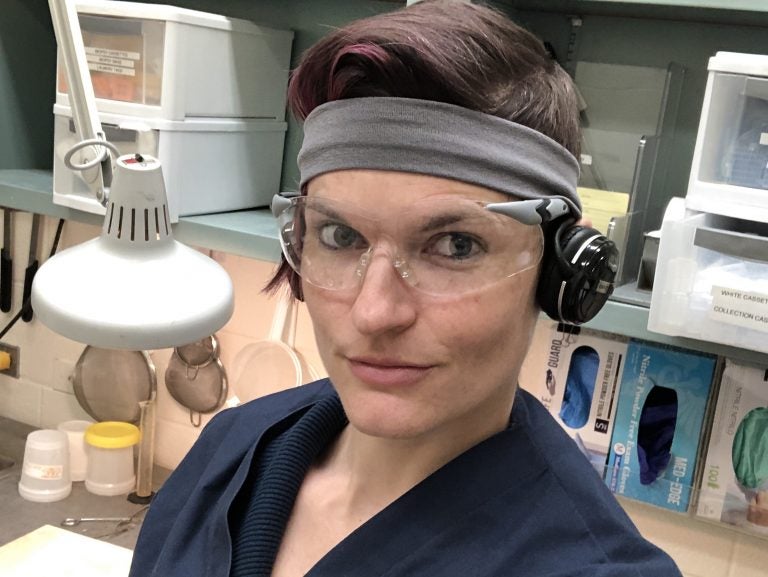 Photo of Kali Holder in her laboratory. Kali is smiling slightly and is wearing scrubs and protective eye glasses.