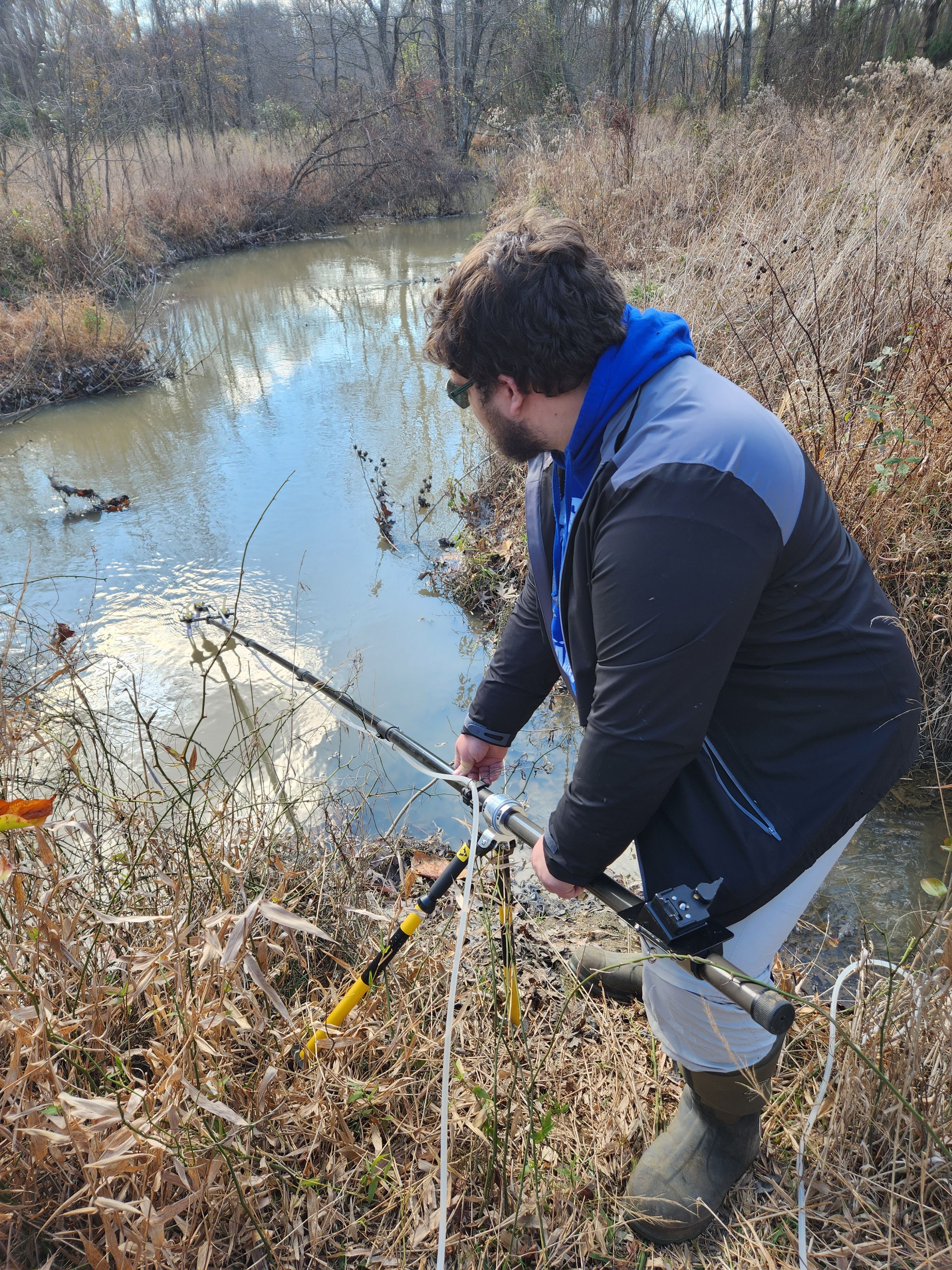 Photo of Sean Lyons taking a water sample. Sean is on the bank of a stream, using a long device on the end of a long pole to collect a water sample.