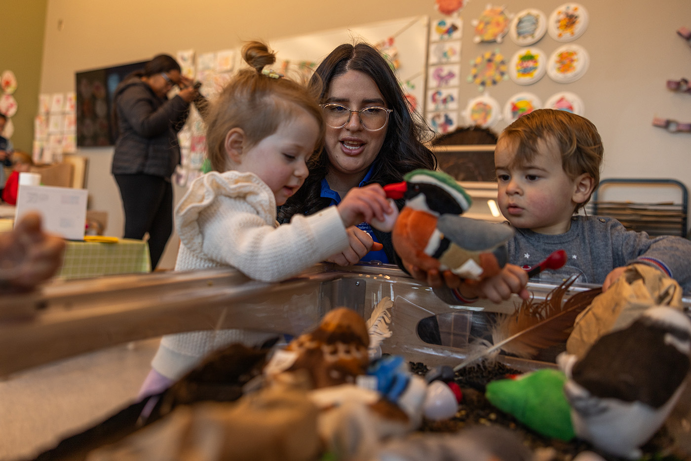 Two children and an educator interact near a tub filled with sand.