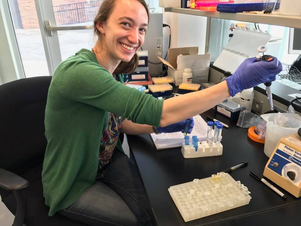 Photo of Alyssa Kaganer in a lab setting using a pipette. She is smiling toward the viewer.