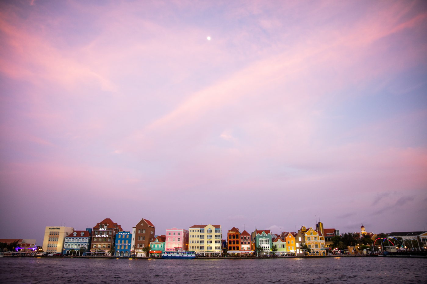 A view from the water of colorful buildings on the shoreline of Curacao under a pink and blue sky during the sunset