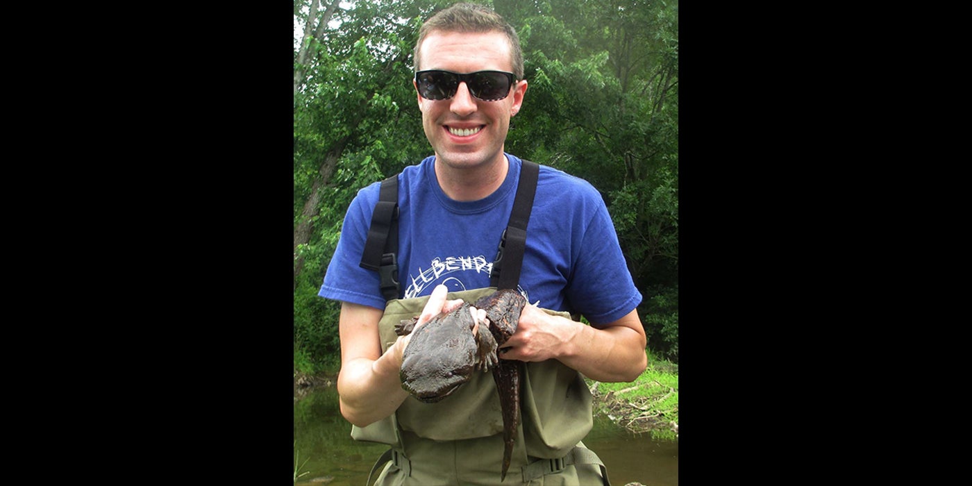 An animal keeper holds a hellbender (large salamander). He is standing outside near trees and a body of water and is wearing sunglasses, a T-shirt and waders.