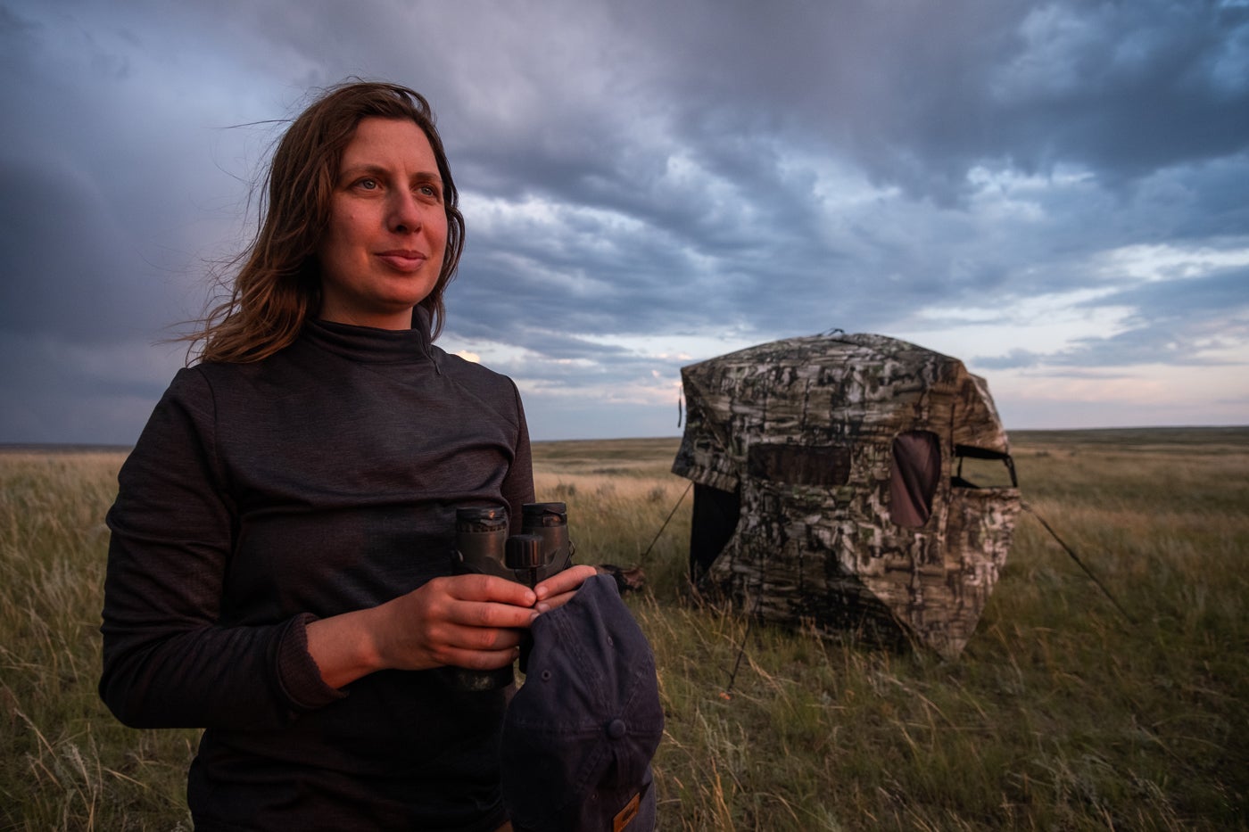 Landscape ecologist Hila Shamon stands on the open prairie in Montana. A camouflage blind tent can be seen behind her, and she holds a hat and a pair of binoculars in her hands.
