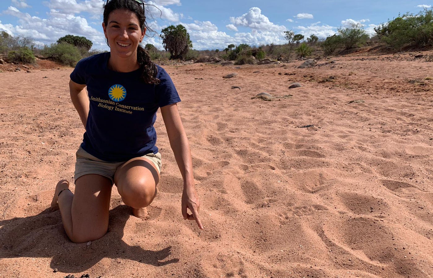 Francesca Vitali takes a knee in a sandy area in Kenya. She's wearing a navy Smithsonian Conservation Biology Institute T-shirt. She points to wildlife footprints in the sand.
