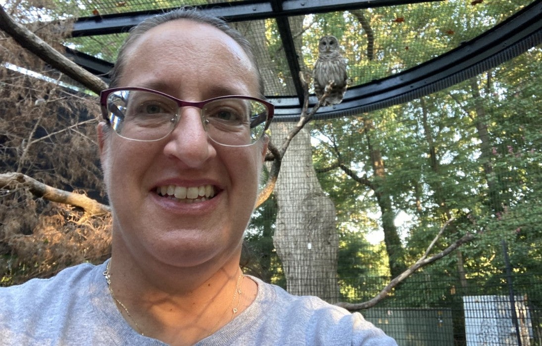 Keeper Heather Anderson takes a selfie in the barred owl yard with male barred owl, Edward, behind her on a tree branch.