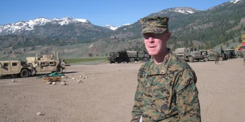 A photo of a young Caucasian man dressed in camouflage fatigues. Other military members and military vehicles can be seen behind him.