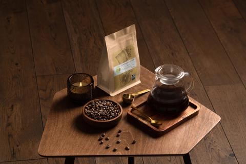 a coffee bag on a table surrounded by coffee beans, a coffee scoop, a pour over, and a candle