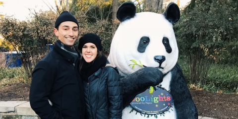 Photo of a smiling young man and woman dressed for winter weather standing next to a larger-than-life sized decorative panda with a Zoolights sign on its chest.