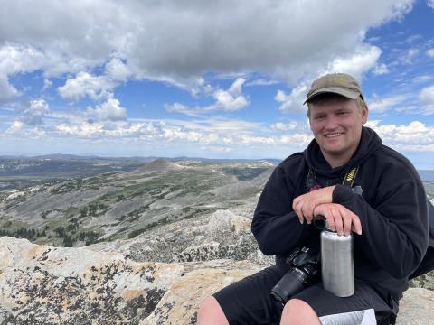 Photo of a young Caucasian man sitting on a mountain top, smiling at the camera.