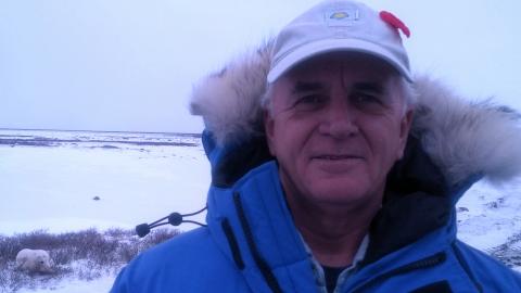 Animal behaviorist Donald Moore wearing a parka and hat in a snowy area where a polar bear can be seen in the background