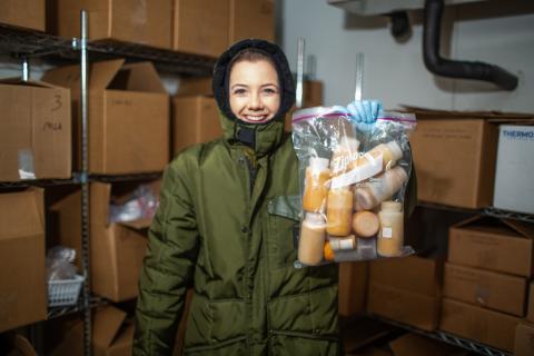 A nutrition lab research assistant at the Zoo wearing a full-body snowsuit stands in the freezer where animal milk samples are stored. She holds a bag full of milk samples, and is surrounded by shelves stacked with cardboard boxes full of more samples.