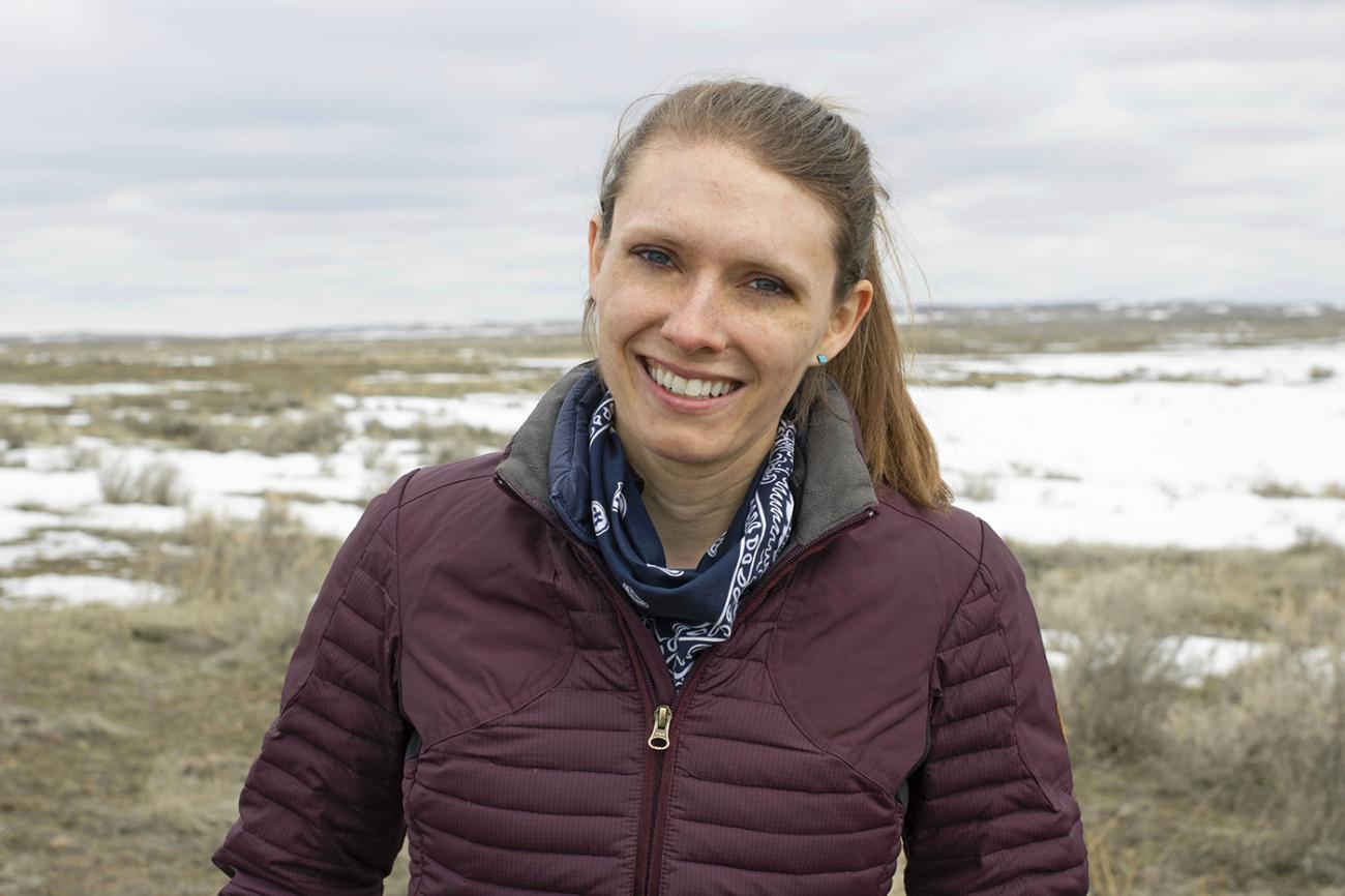 A light haired woman in a puffy maroon coat smiles in front of a snowy prairie.