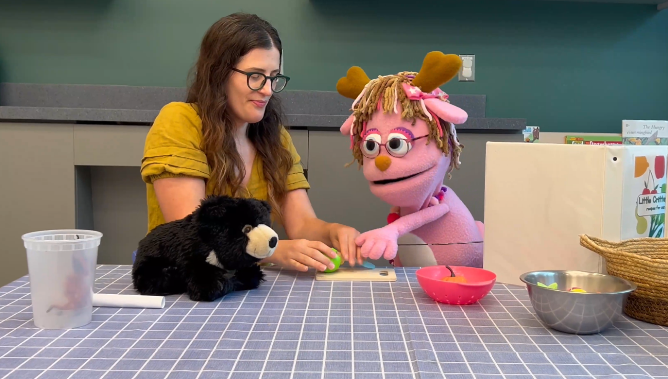 A teacher helps a pink jackalope puppet use a toy knife to chop a toy apple to feed to a stuffed sloth bear