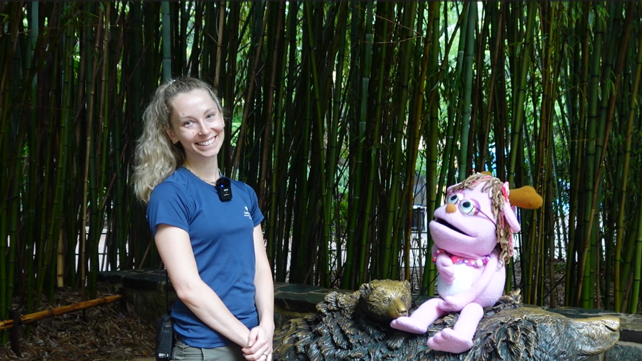 A woman stands next to a pink puppet with green foliage in the background.