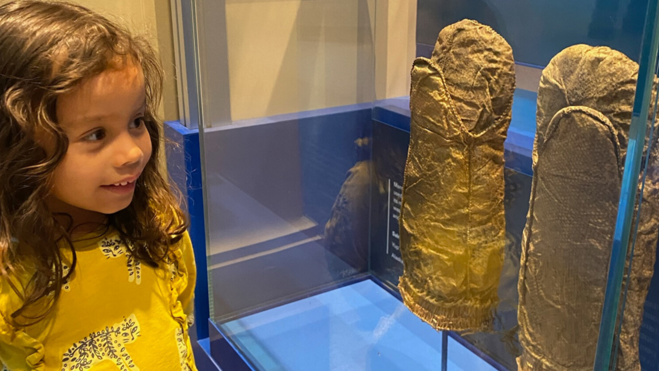 A young child with an interested expression on her face looks at a pair of large fish skin mittens crafted by one of the indigenous nations of Northern North America