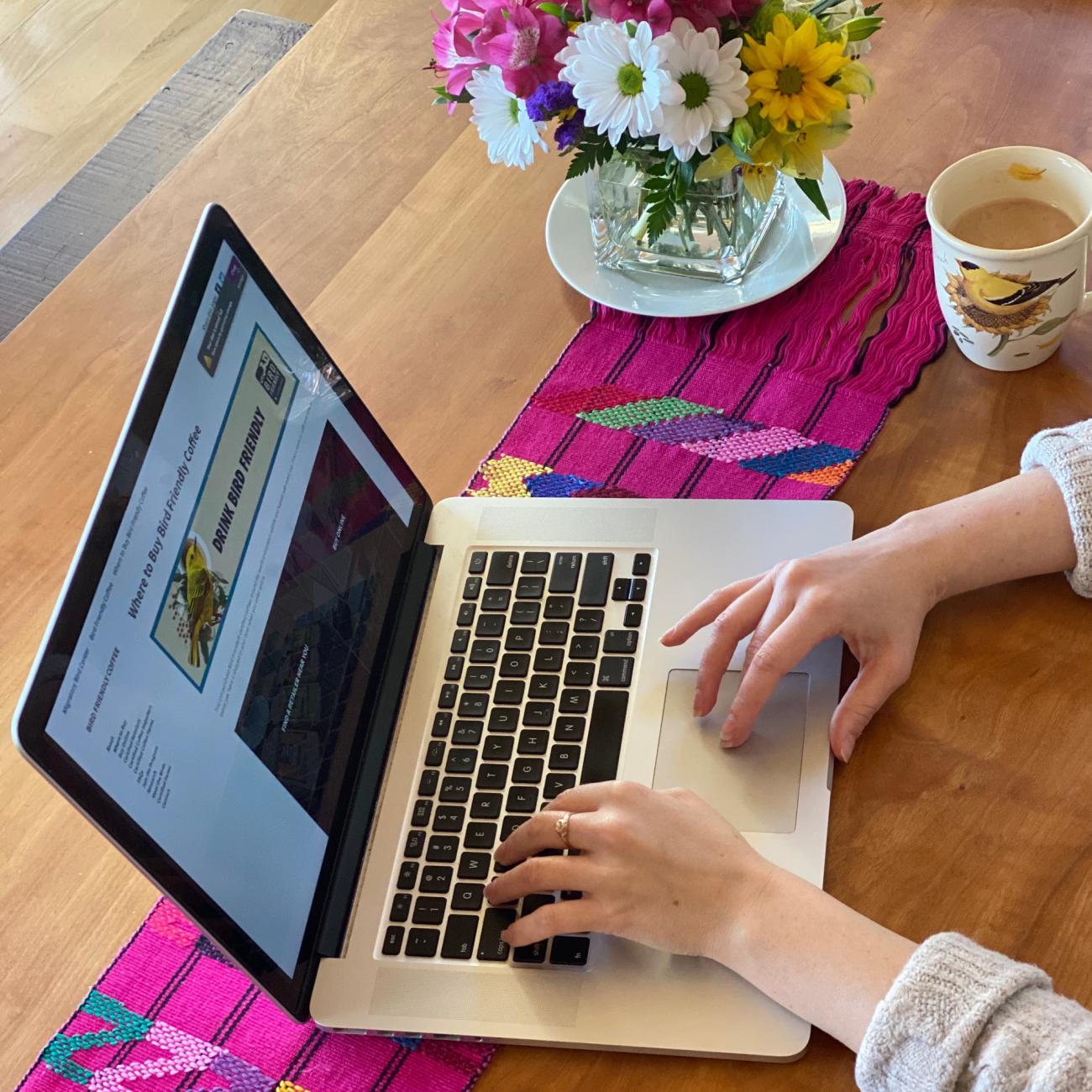 A photo of a person using a laptop. A mug of coffee and a flower base are also on the table.