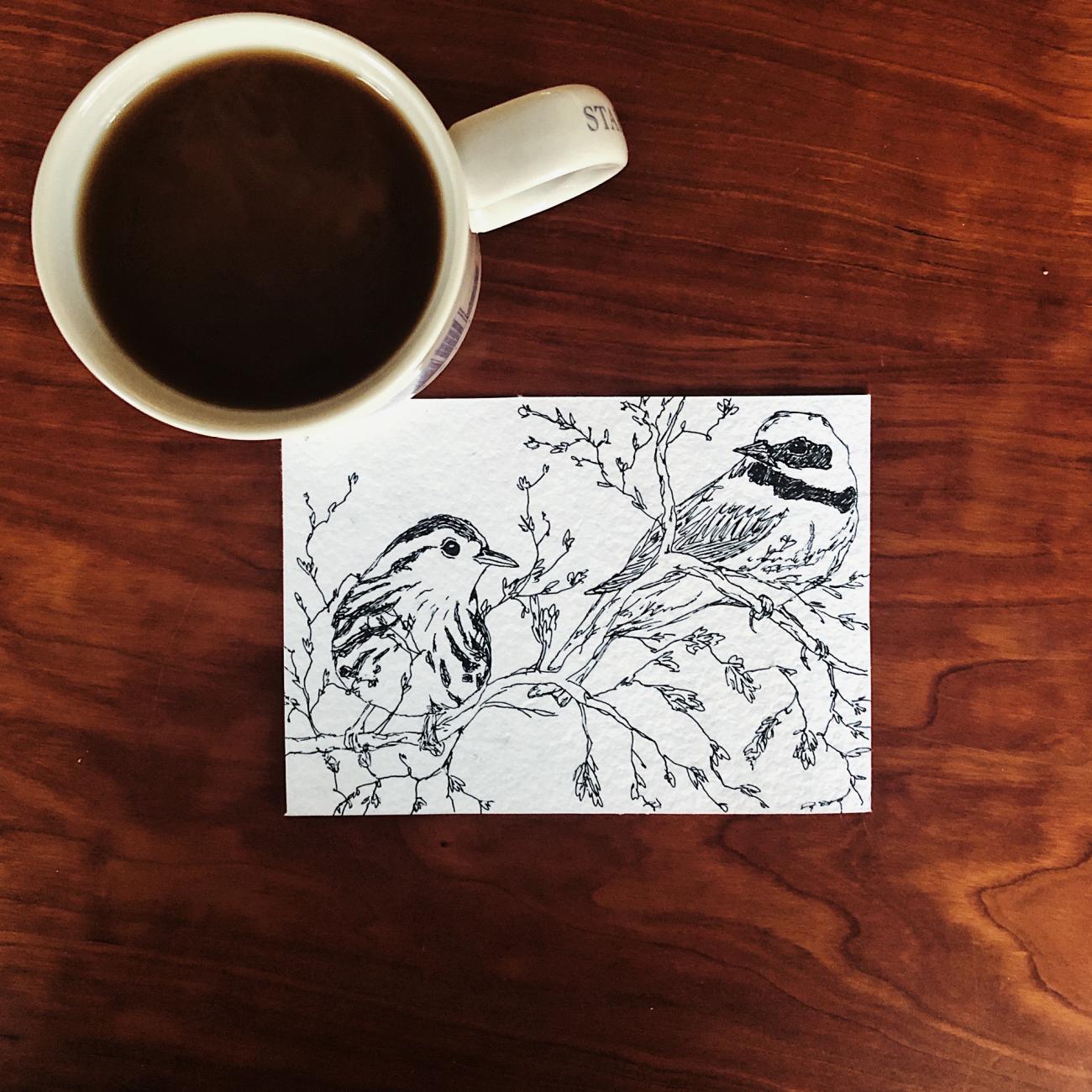  A mug of coffee placed next to a pen and ink drawing of two migratory birds perched on a tree branch