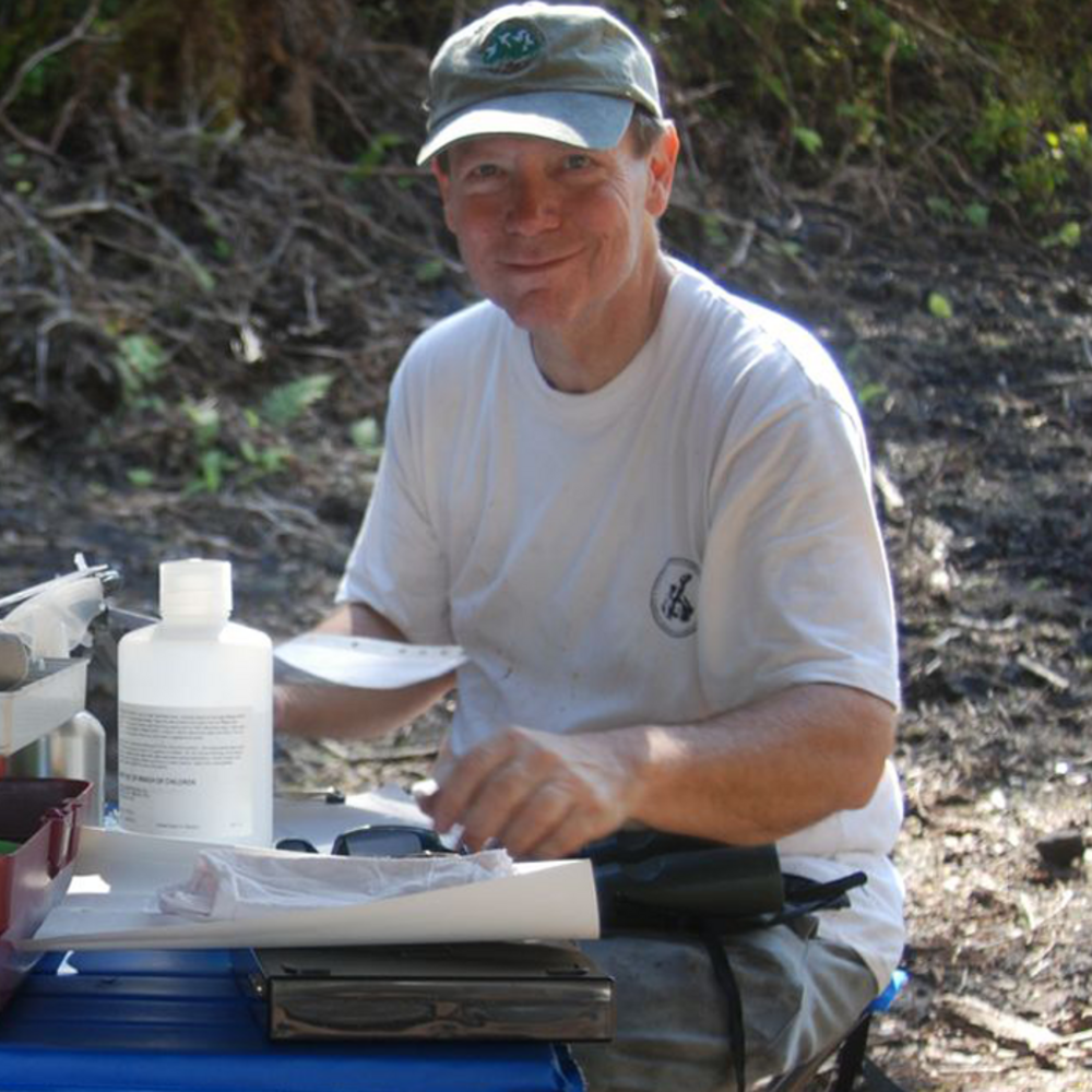 Portrait of a man wearing a white t-shirt and a blue baseball cap sits at a desk filled with scientific equipment in the Hawaiian jungle.
