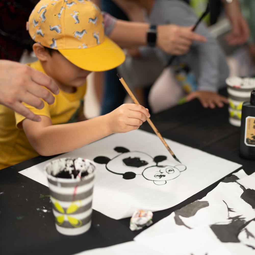 A child wearing a yellow hat draws a panda with a calligraphy brush.