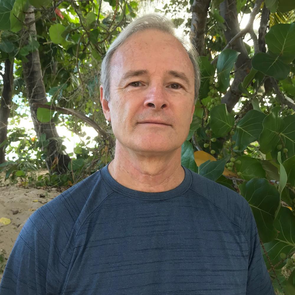 A white-haired scientist poses for a headshot with tropical trees behind him.