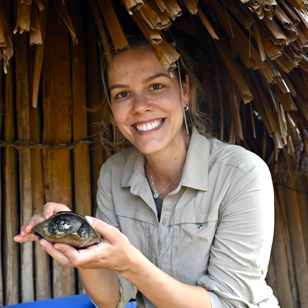 A female scientist wearing a beige field shirt poses in front of a wicker building while holding a small, dark-colored turtle