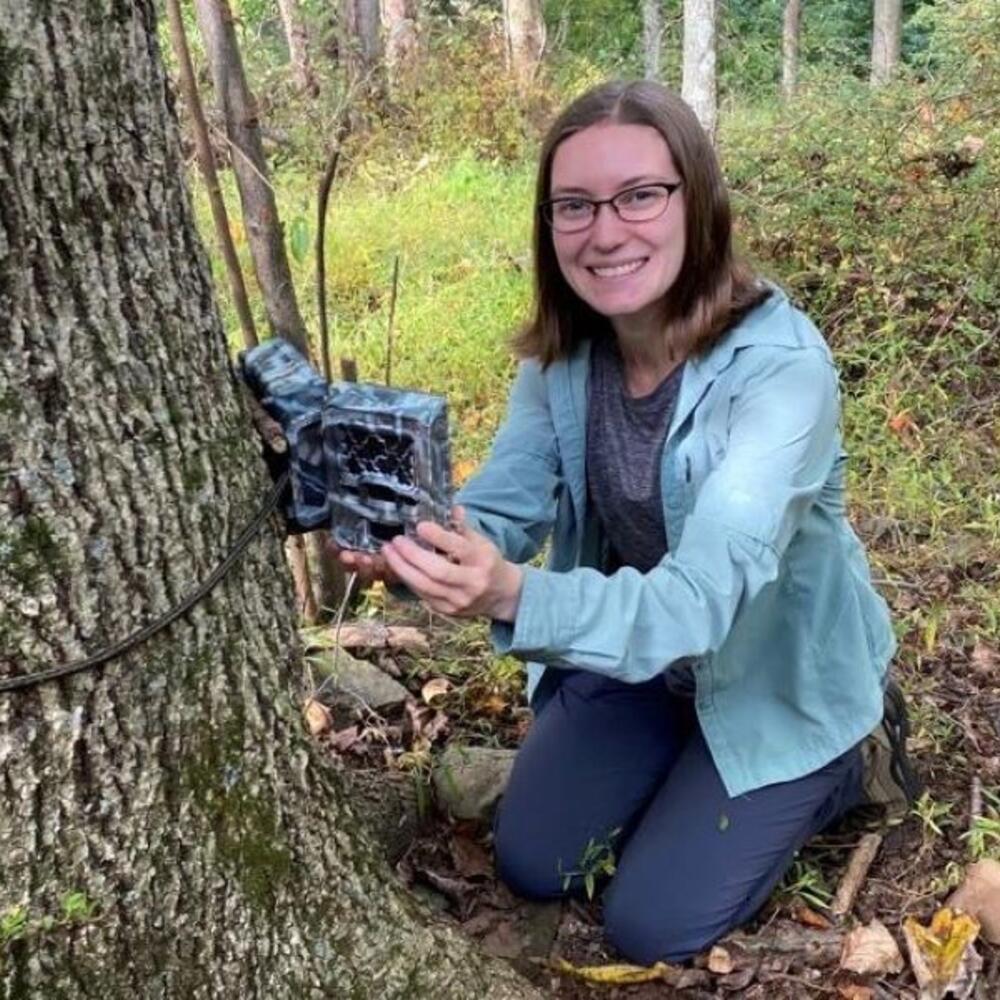 A female scientist posing next to a camera trap tied to a tree