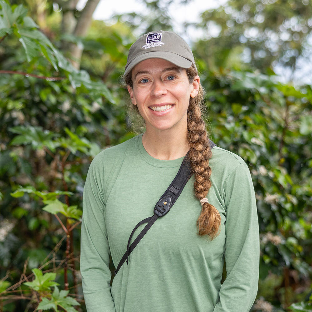 A female scientist wearing a baseball cap stands in front of a leafy background.