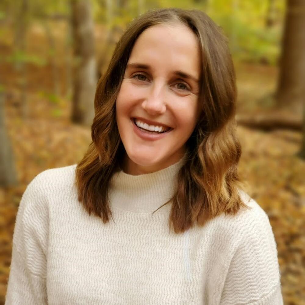 A smiling researcher with long brown hair and a cream-colored sweater is seen in a forest.