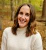 A smiling researcher with long brown hair and a cream-colored sweater is seen in a forest.