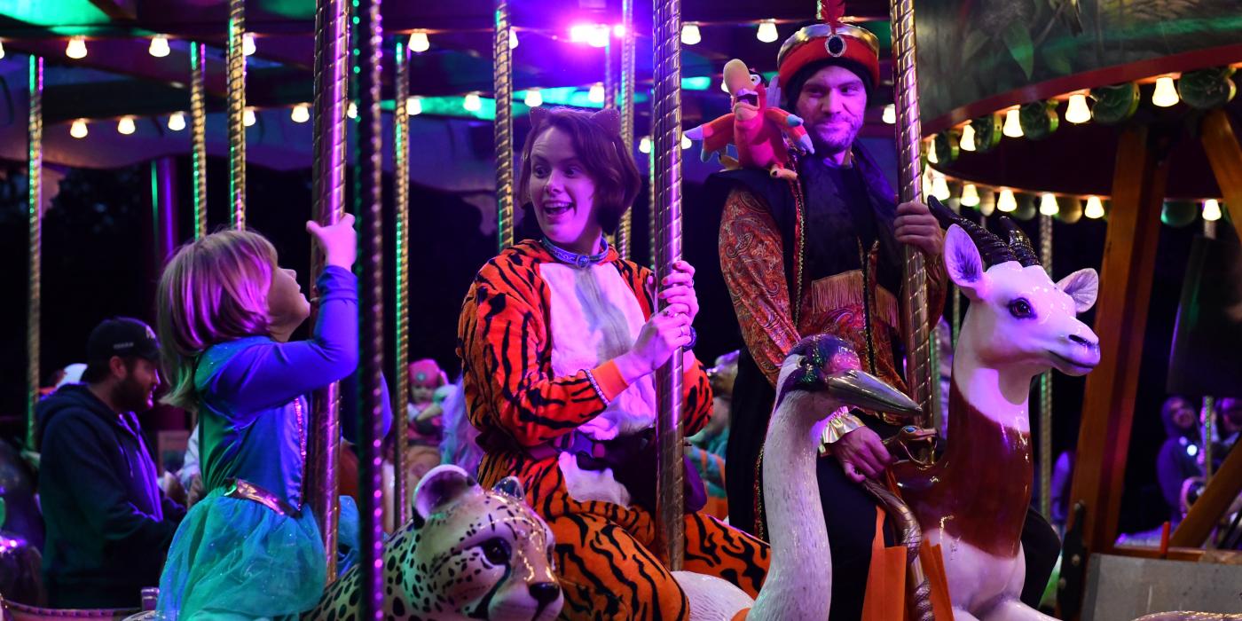 Photo of a family of two adults and a young child riding a carousel together. All three people are wearing Halloween costumes.