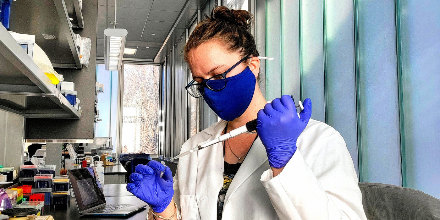 Female scientist wearing a PPE mask and lab coat using a pipette in a lab.