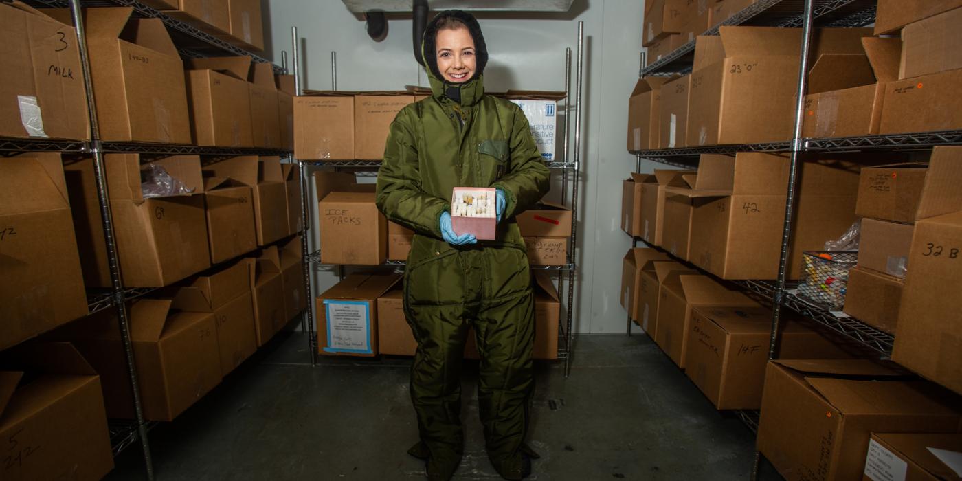 A nutrition lab research assistant at the Zoo wearing a full-body snowsuit stands in the freezer where animal milk samples are stored. The shelves are stacked with cardboard boxes and she holds a small box filled with tubes of milk samples.