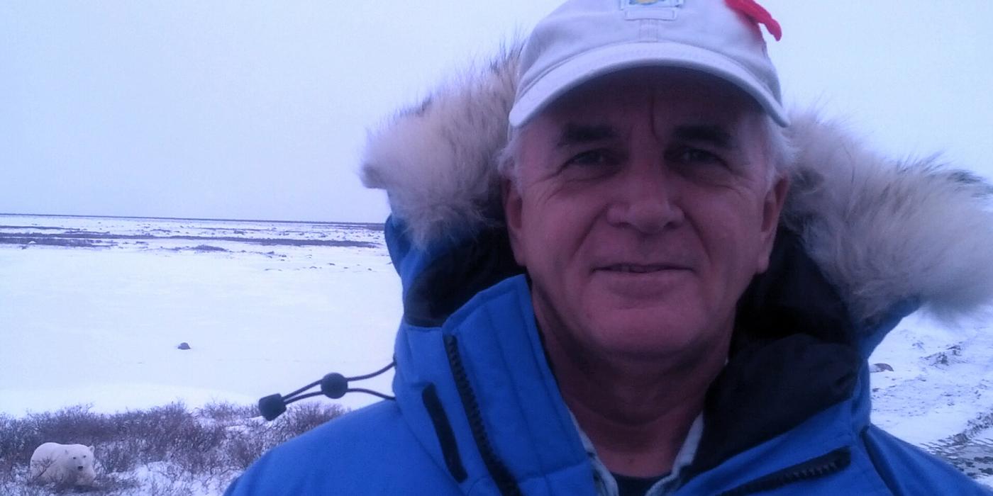 Animal behaviorist Donald Moore wearing a parka and hat in a snowy area where a polar bear can be seen in the background