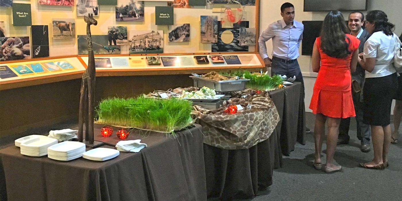 A catered event in the Smithsonian's National Zoo's Think Tank exhibit. A group of people stand and chat near a set of tables with black table cloths, plates and catered food. Exhibit panels can be seen in the background.